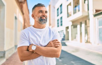 Man with a beard wearing a white t-shirt stands outdoors on a sunny street, arms crossed, looking to the side.