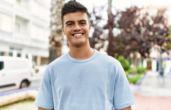 A person smiling outdoors, wearing a light blue T-shirt. Blurred background includes trees and buildings.