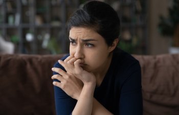 A person with dark hair looks concerned, sitting on a brown couch with their hand covering their mouth. Background shows a blurry bookshelf.