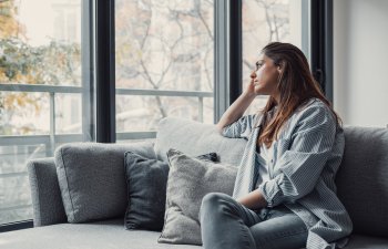 Woman sitting on a couch, looking pensively out a large window. She rests her chin on her hand, with trees visible outside.