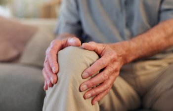 Senior, man and hands for knee pain in home, arthritis and osteoporosis and leg issue for healthcare. Living room, fibromyalgia and sore joint on couch in lounge, injured and emergency problem