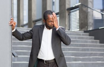 An African-American man in a suit feels bad, dizzy and has a headache, loses consciousness, closes his eyes, holds his hand to his head, needs help.