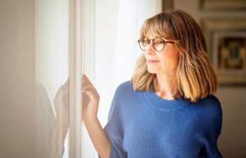 Portrait of a blond haired mid aged woman looking out of the window and smiling. Confident female wearing eyeglasses and blue shirt.
